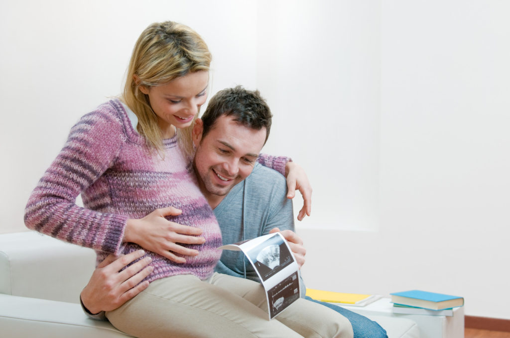 Young smiling couple holding and looking at an ultrasound scan of their expecting baby at home, copy space